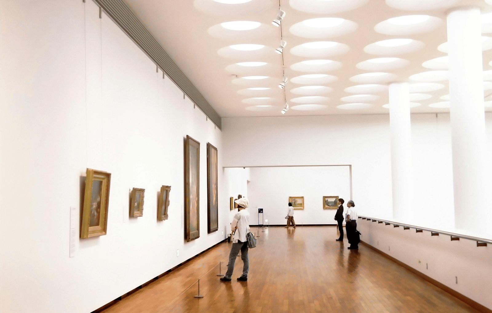 A spacious and elegantly designed museum gallery interior featuring high ceilings, polished wooden flooring, and framed artworks displayed along the walls, with visitors browsing the expansive exhibition space.