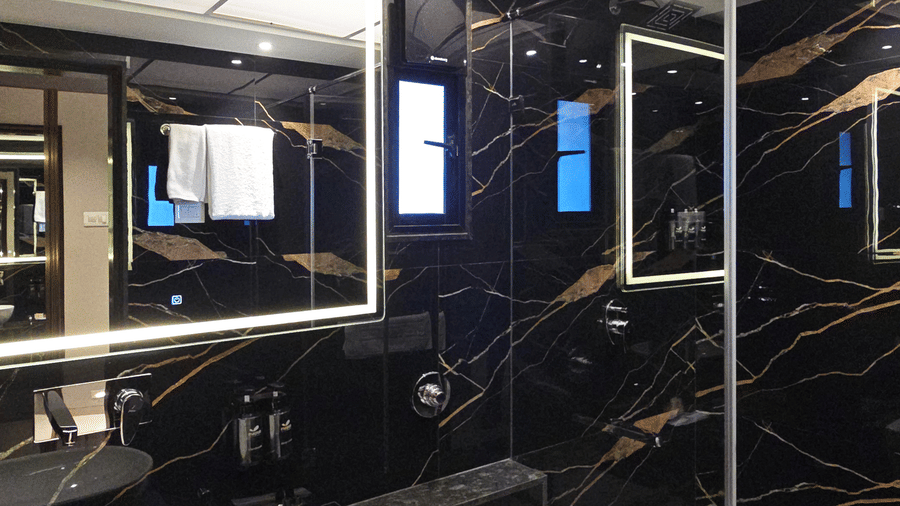 A black marble bathroom showing a lit vanity mirror, a vessel sink, a wall-mounted toilet, and a glass-enclosed shower area at Magnus Kapitol, Sakore Nagar, Viman Nagar.