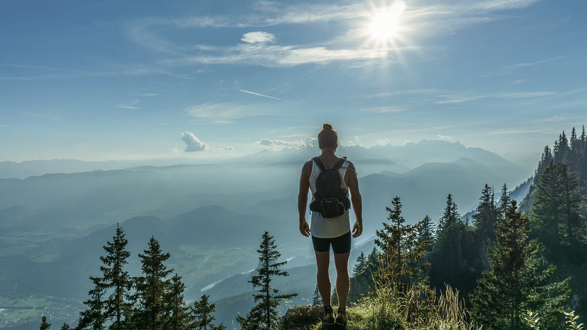 A person standing on a hilltop overlooking layered mountain ranges and trees under a bright sky.
