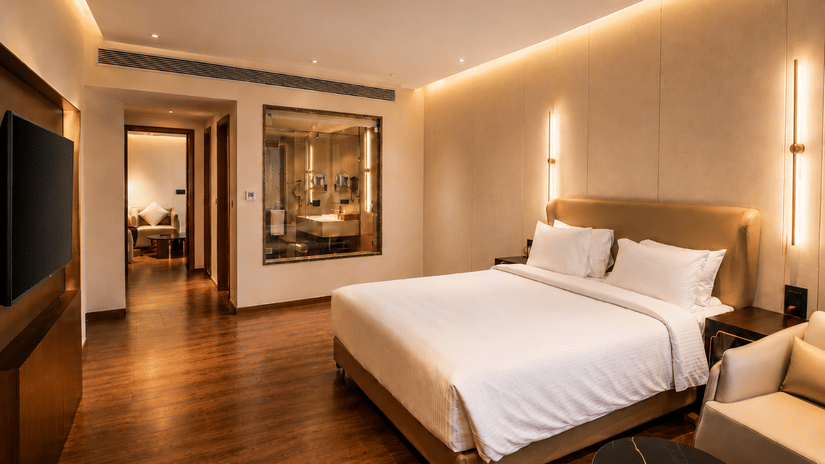 A Premium Room at Pride Plaza Indore featuring a bed with pillows, wall sconce lighting, a television wall panel, and an entryway to the bathroom.