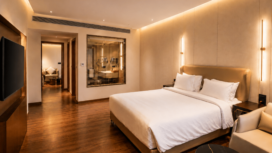 A Premium Room at Pride Plaza Indore featuring a bed with pillows, wall sconce lighting, a television wall panel, and an entryway to the bathroom.