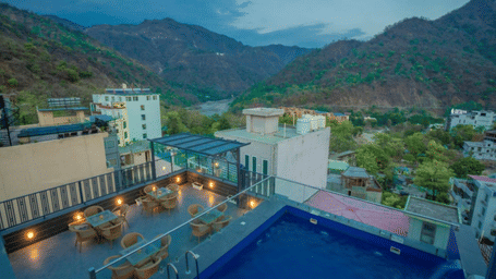 An aerial view of Perfectstayz Premium Laxmi Heritage, Rishikesh property with a swimming pool, surrounding buildings, and hills in the background captured during evening light.