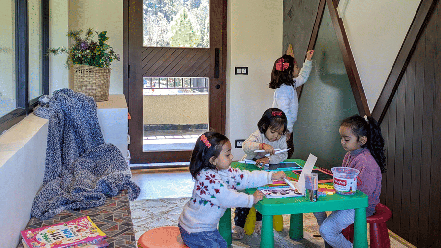 4 children drawing at a green table with coloured stools while one writes on a chalkboard wall, with picture books on the bench at The Chamomile Estate.