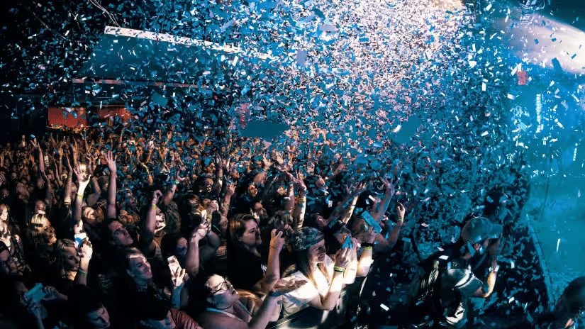 A large crowd of people with hands raised at an indoor party event with confetti falling from above and colorful stage lights illuminating the venue in the background.