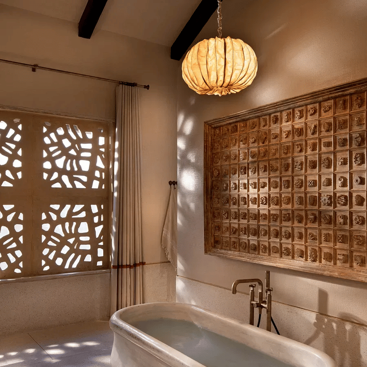 A bathroom with a freestanding bathtub positioned near a window with a patterned screen, casting shadows on the wall. A round, woven pendant light illuminates the space. A section of patterned tiling or screen is visible above the bathtub.