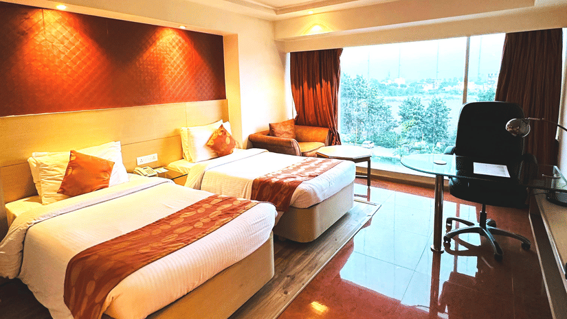  A hotel room at Amer Greens featuring two beds with orange bed runners, a dark headboard, and a large window overlooking the outdoors.