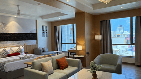 Wide view of the Club Room at Lords Inn Somnath highlighting the king-sized bed, comfortable sofa seating area, and bright window view.