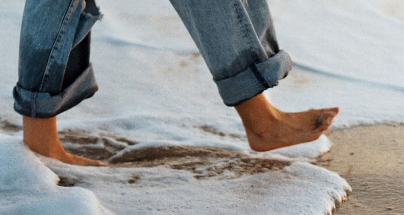 a person wearing baggy denim pants walking on the beach with gentle waves crashing on his feet
