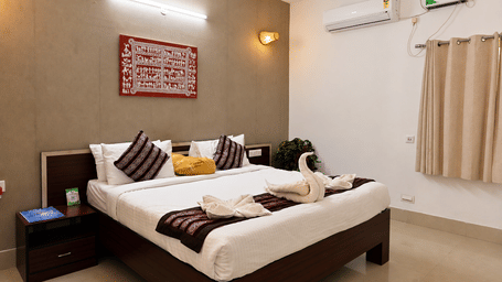 Hotel room with a bed dressed in white linen, swan-shaped towel art, patterned cushions, a wall-mounted artwork and a window with curtains.