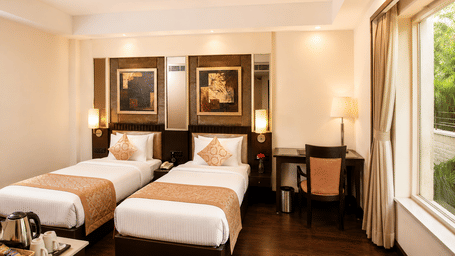 Hotel room with twin beds dressed in white linen, framed wall artwork, a work desk and long curtains by a window overlooking green trees.
