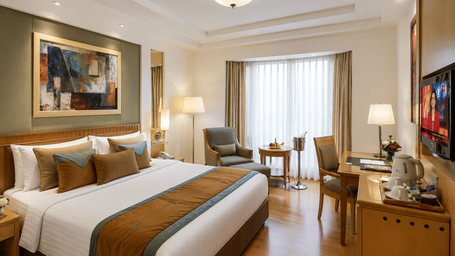Hotel room with a bed dressed in white and gold linen, a wall-mounted artwork, a seating area with an armchair and a wooden work desk nearby.