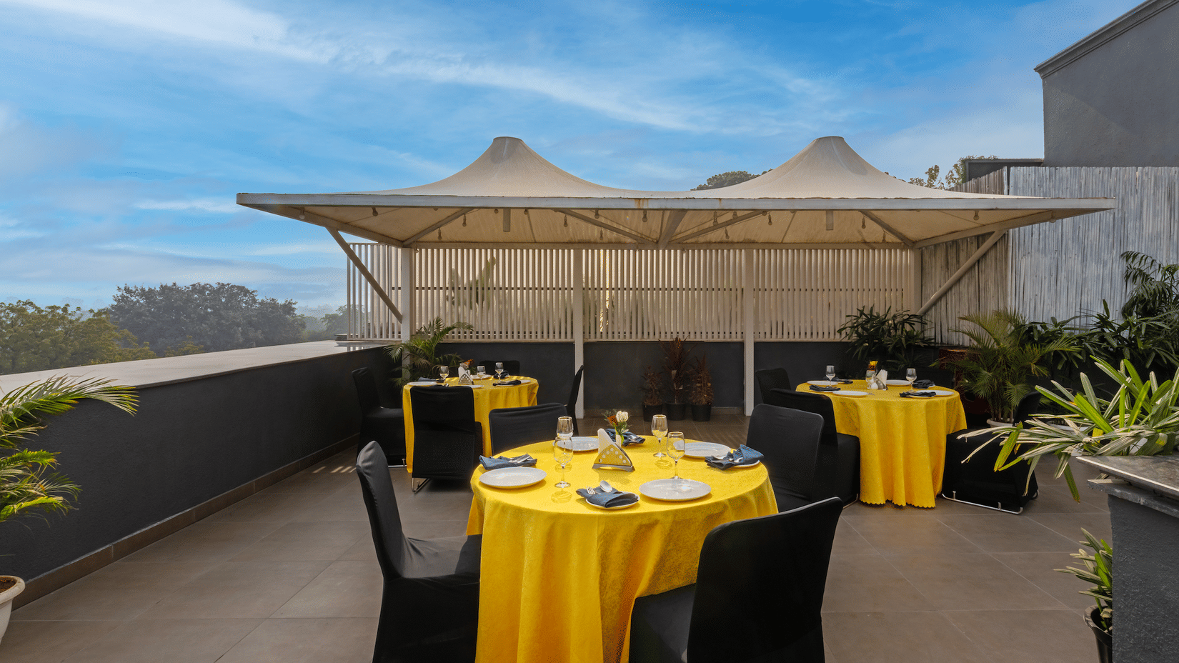 Open terrace dining setup with a canopy, rectangular table with yellow covers, and four chairs at Hotel Saket 27, New Delhi.