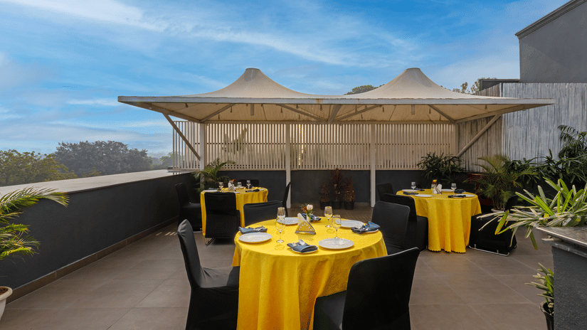Open terrace dining setup with a canopy, rectangular table with yellow covers, and four chairs at Hotel Saket 27, New Delhi.