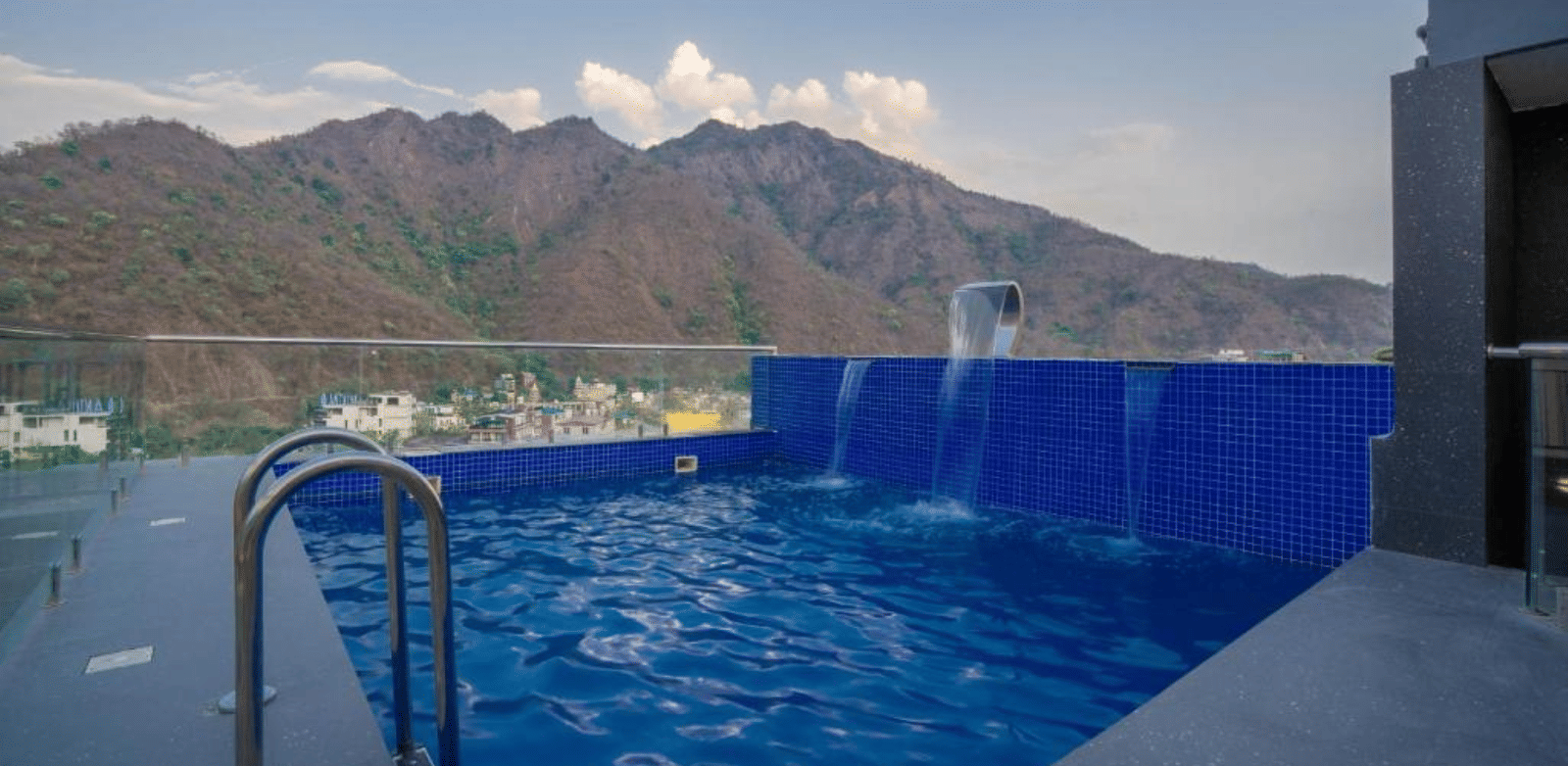 An outdoor swimming pool with metal handrails, water flowing from a spout, and a mountain range visible in the background under a clear sky at Perfectstayz Premium Laxmi Heritage.