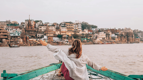 A person with long hair sitting on the bow of a boat with arms outstretched, facing a city skyline along a river.