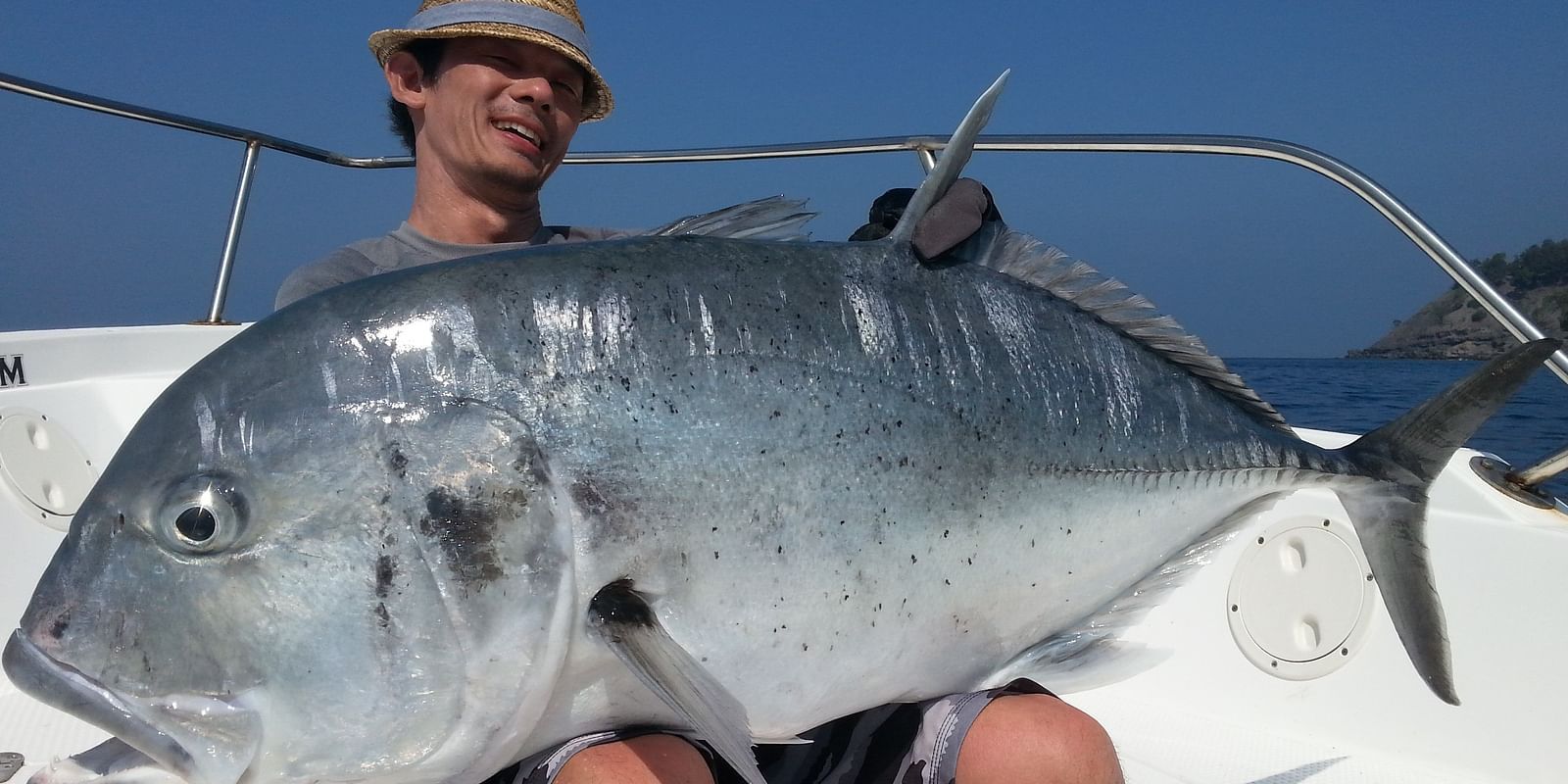 A man sitting on a boat, holding a large fish resting on his thighs.