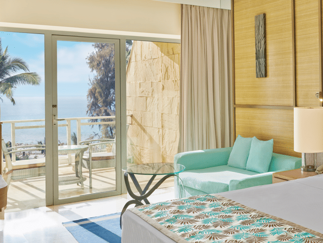 A bedroom with a king size bed, mint-green sofa, glass coffee table, and a balcony offering a clear view of the sea and palm trees at The Resort, Mumbai.