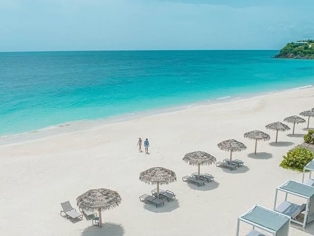 White sandy beach with loungers and cabanas at Tamarind Hills Resort and Villas, overlooking the turquoise Antigua Sea under a clear blue sky.