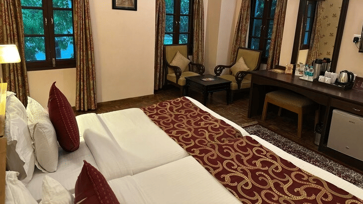 A bedroom with twin beds joined together, 2 armchairs by the window, and a wooden vanity desk at Amantra Shilpi Resort, Udaipur.