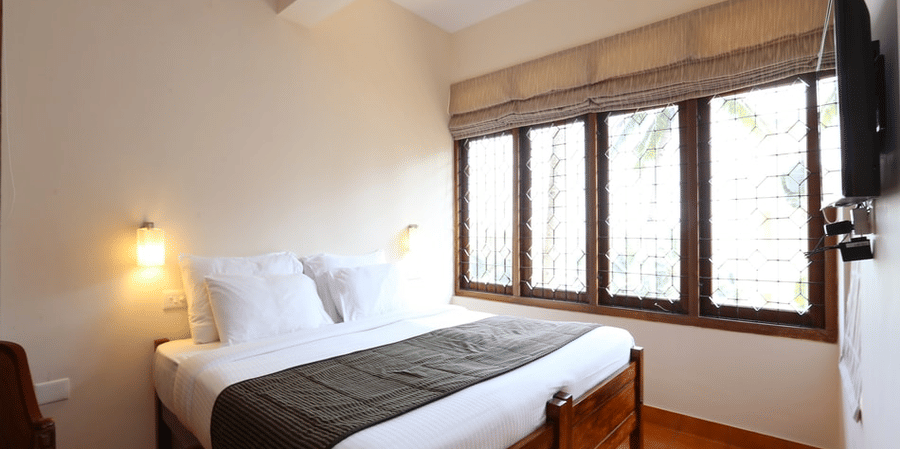A deluxe room bedroom at Abad Serviced Villas, Kovalam with a double bed, bedside table, lamp, and large windows.
