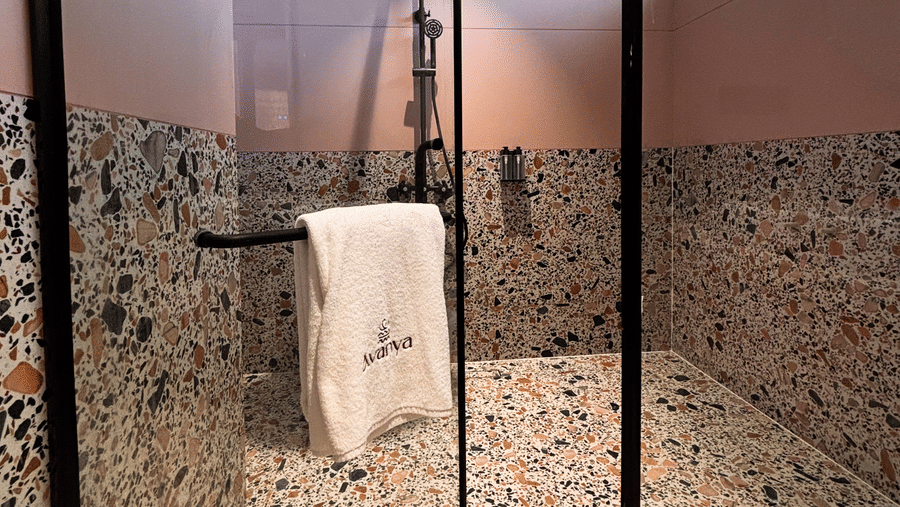 A glass-enclosed shower area features multi-coloured terrazzo wall and floor tiles with a white towel on the handle at Avanya Resorts N Retreat.