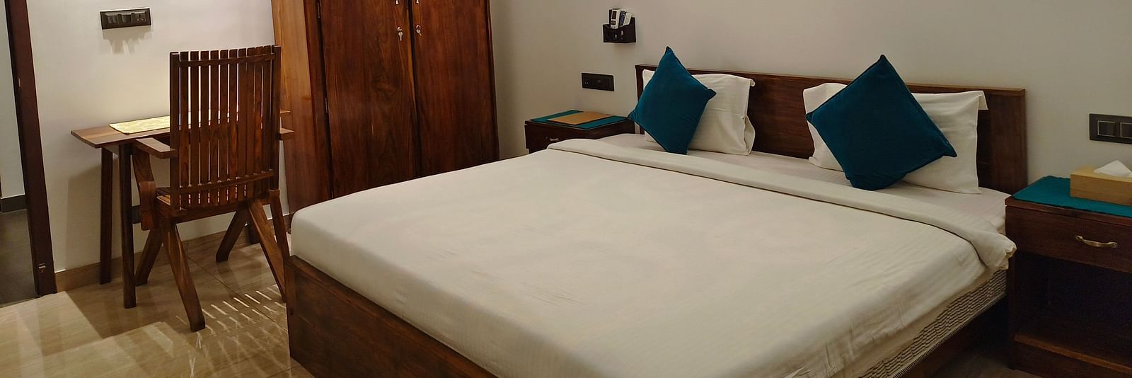 Wide view of a double bed with blue pillows, a chair and a cupboard in 102 room at Ananda Holiday Home, Wayanad 9