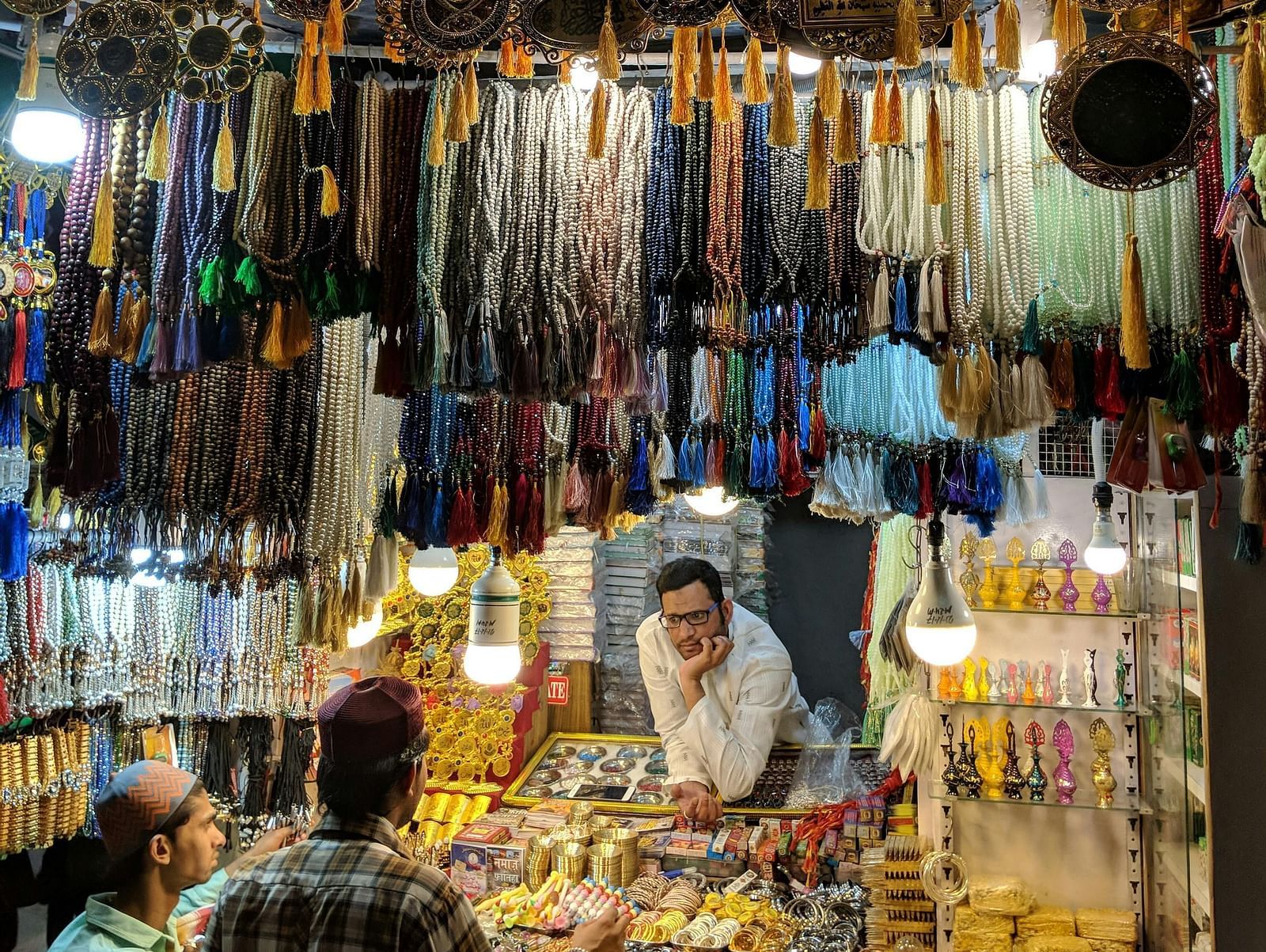 A bustling traditional market stall selling vibrant bangles, decorative items, and colourful accessories, illuminated by warm lighting.