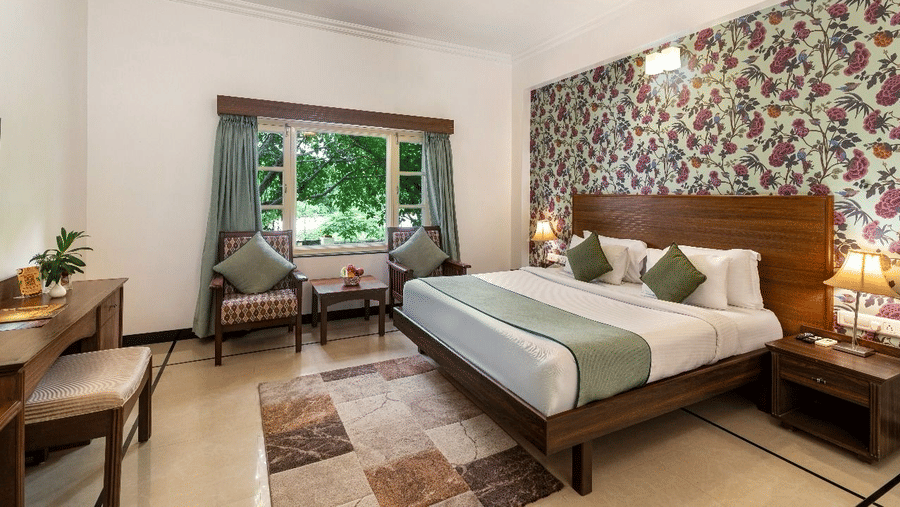 A guest room featuring a king size bed, a floral wallpaper accent wall, 2 armchairs by a window, and a desk at Amantra Shilpi Resort, Udaipur.