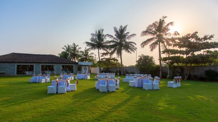An outdoor event setup on a green lawn with round tables and chairs, and a building with palm trees in the background, under evening light  | Grande Pavillion | Grand Bay Resort and Spa