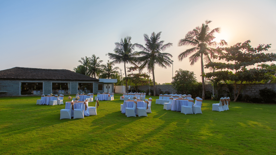 An outdoor event setup on a green lawn with round tables and chairs, and a building with palm trees in the background, under evening light - Grande Bay Resort & Spa, Mamallapuram