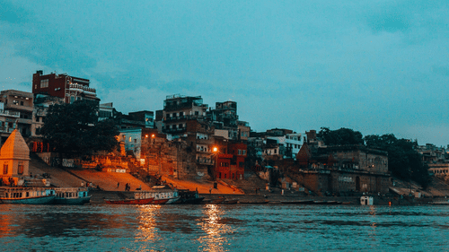 A view of the Ganges River at dusk with illuminated buildings along the Varanasi ghats.