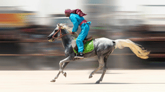 A rider in blue clothing and a red headscarf gallops a gray horse across a track with a heavy motion blur effect.