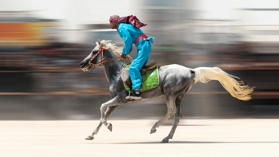 A rider in blue clothing and a red headscarf gallops a gray horse across a track with a heavy motion blur effect.