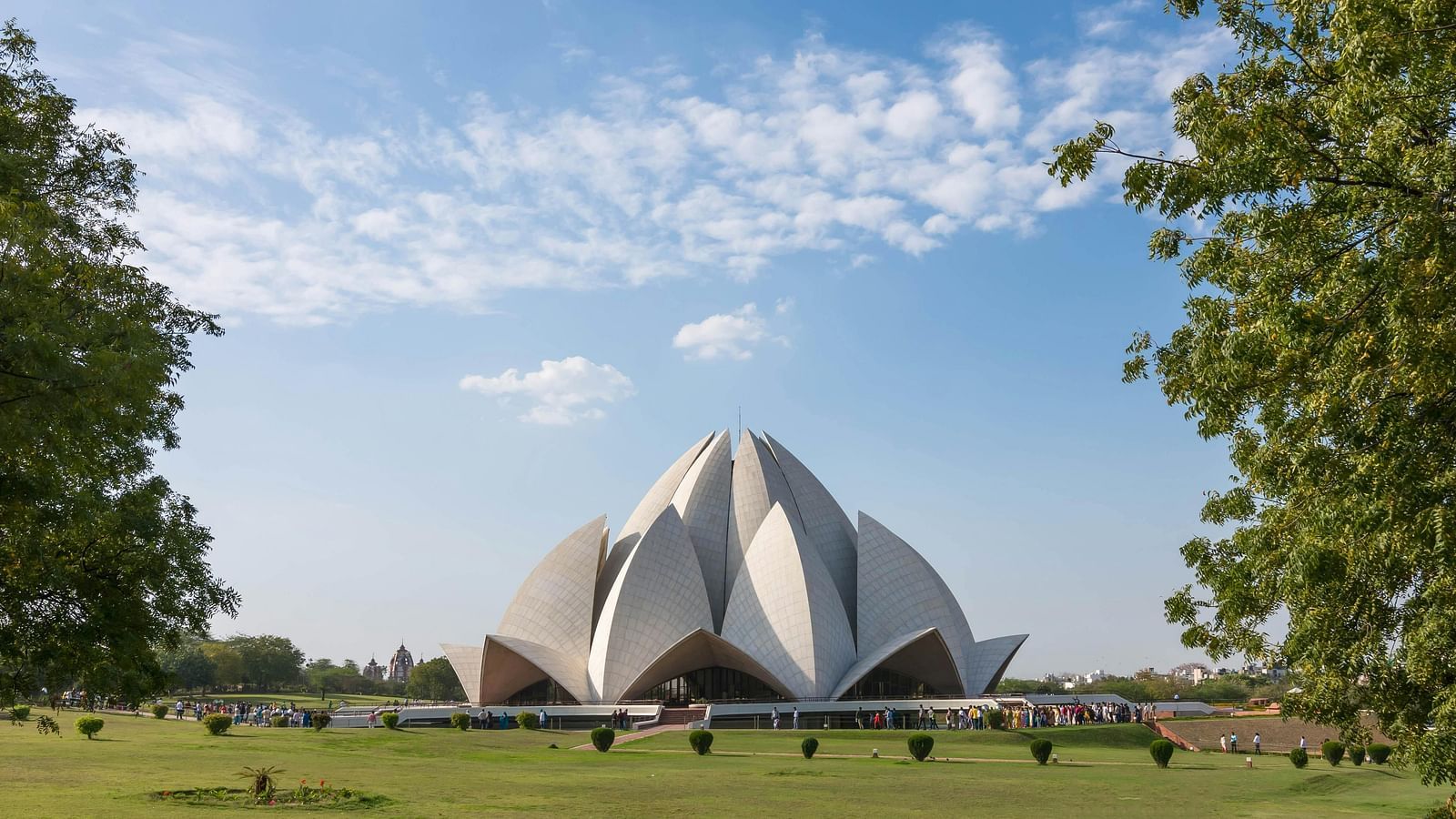 The Lotus Temple, a modern architectural marvel shaped like a lotus flower with white petal-like structures, stands in a green landscape under a blue sky with scattered clouds.