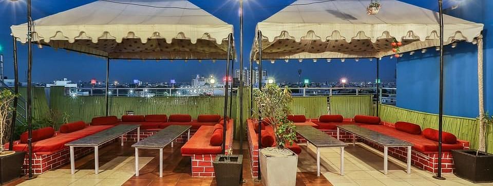 A twilight view of a rooftop dining space with long tables, red cushioned seating and an urban backdrop - TGI Apple Inn, Jaipur 