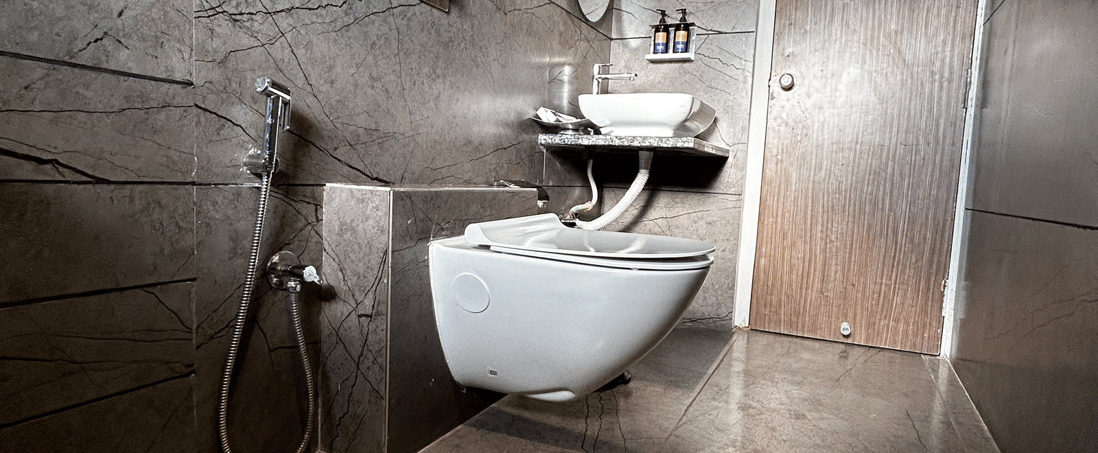 A contemporary hotel washroom with a wall-mounted toilet, modern sink, and dark wood-effect tiled walls at Pravasa Hotel, Kolhapur.