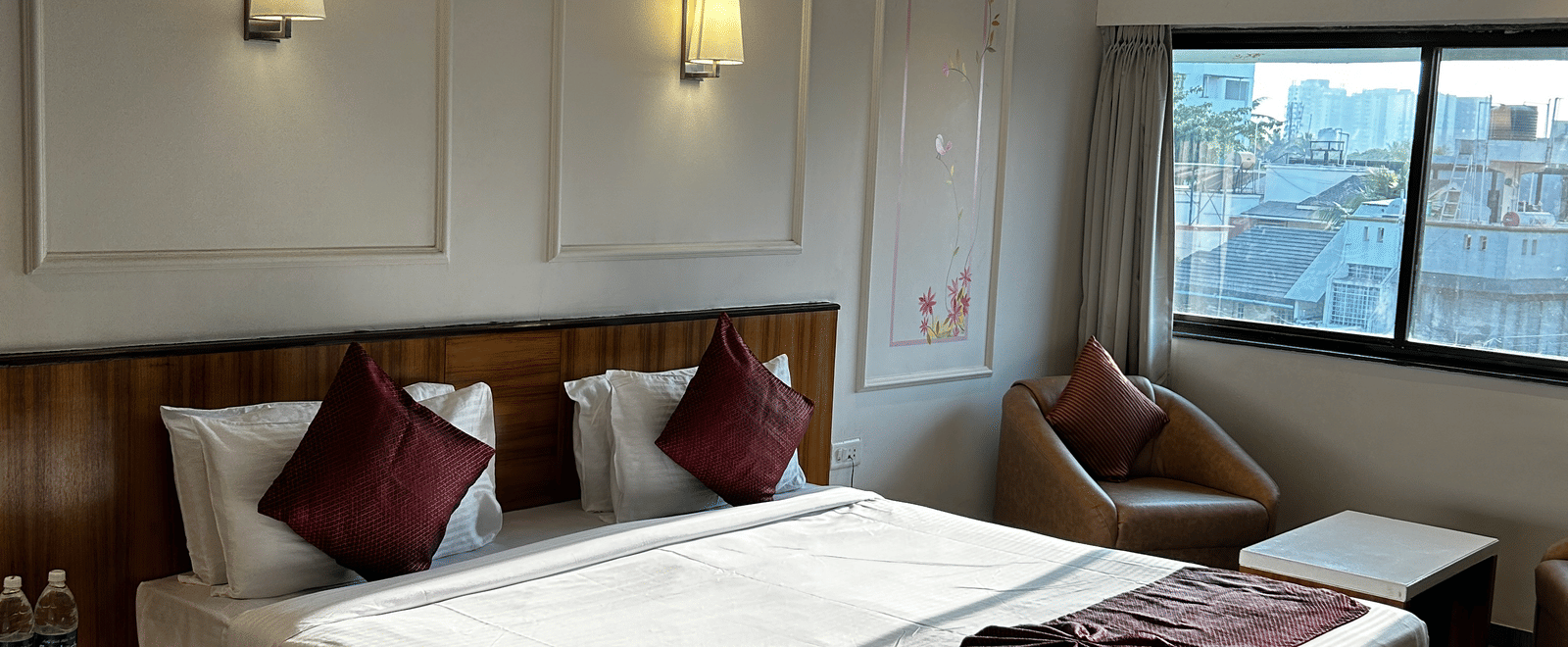  A comfortable guest room at Pravasa Hotel, Kolhapur, featuring a double bed, subtle wall panel moulding, and a wicker armchair.