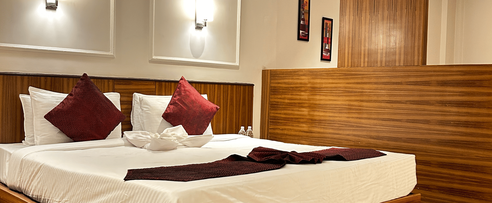 A contemporary double bedroom with a wooden headboard, red throw pillows, and a partial wooden room divider at Pravasa Hotel, Kolhapur.