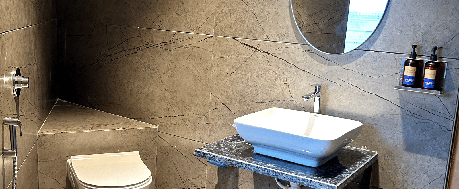 A contemporary washroom with slate-grey tiled walls, a vessel sink, and a modern circular mirror at Pravasa Hotel, Kolhapur.