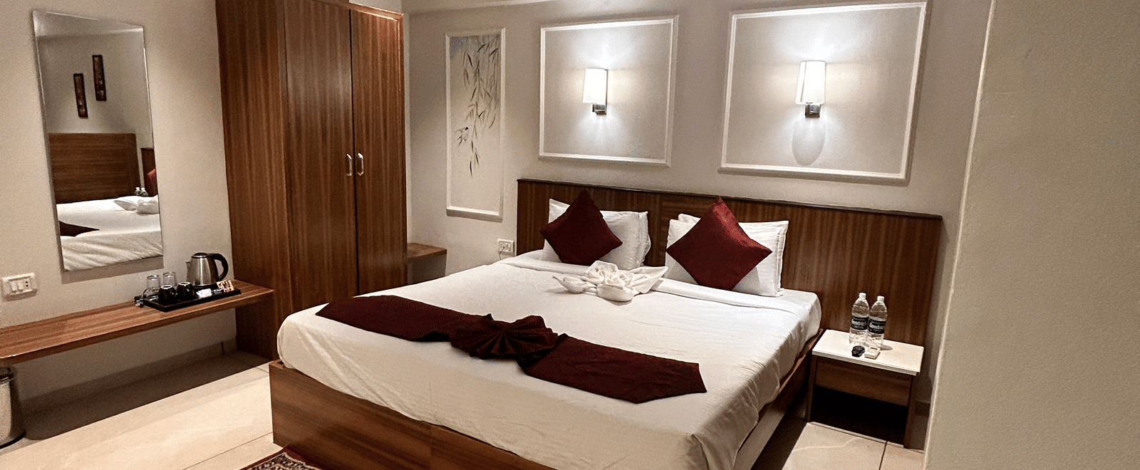 A warm, well-lit standard bedroom featuring a double bed, wooden furniture, and a ceiling fan at Pravasa Hotel, Kolhapur.