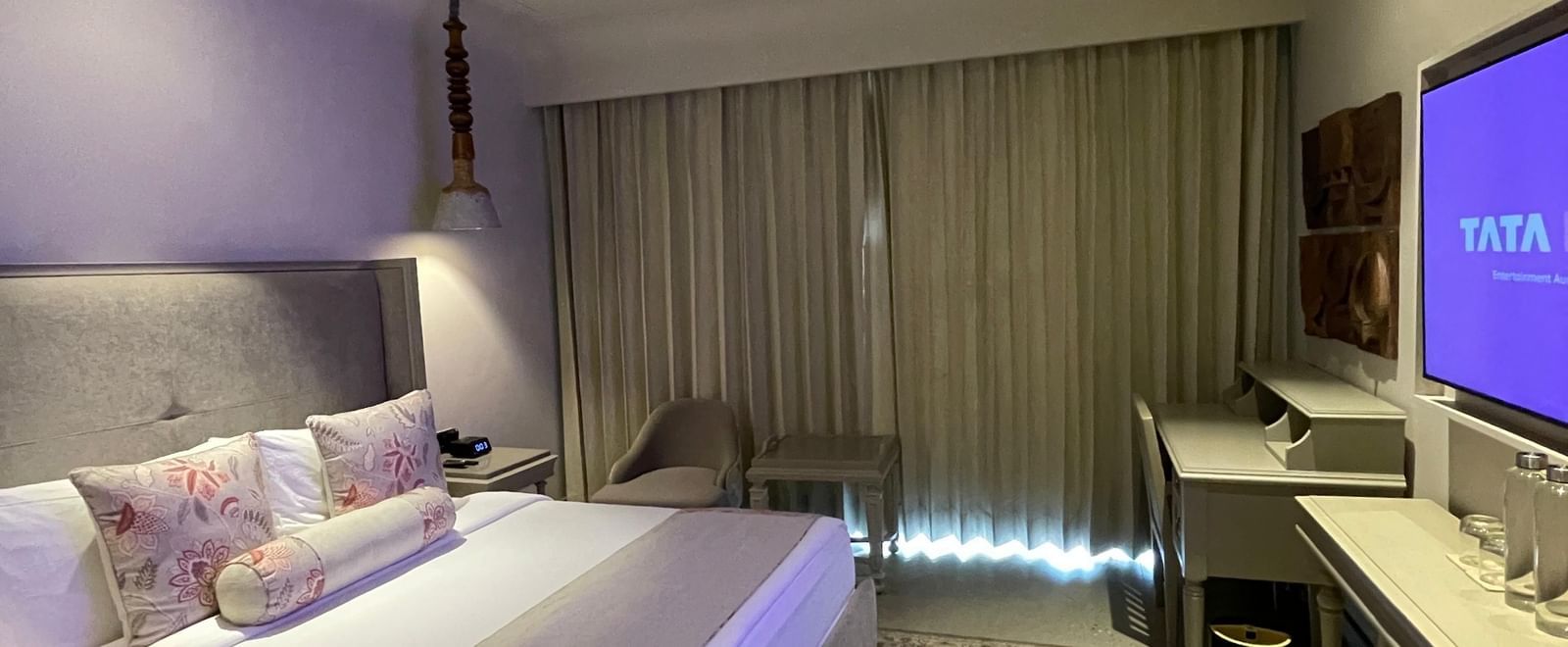 A view of the spacious bedroom at The Evren, Vagator, with a large bed featuring decorative pillows and a bed runner. The room has a patterned rug, a desk, a mounted television, and a window with curtains.