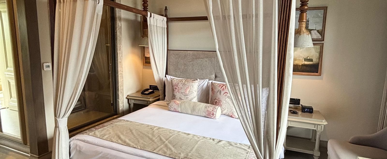 A wide shot of a Premium King Room at The Evren, Vagator, featuring a four-poster bed with elegant curtains and a patterned rug. The room has a high, sloped wooden ceiling and an open doorway leading to the bathroom.
