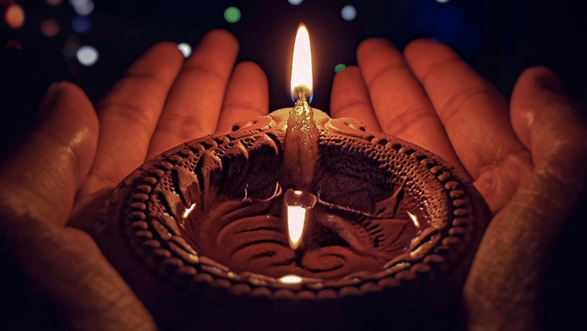 Hand holding a traditional clay lamp with colourful lights glowing softly in the background during Karthigai Deepam
