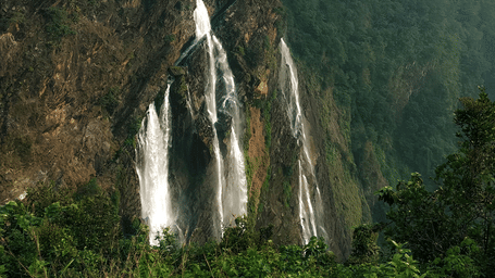 A tall waterfall flowing down a steep rocky cliff surrounded by dense green forest foliage.