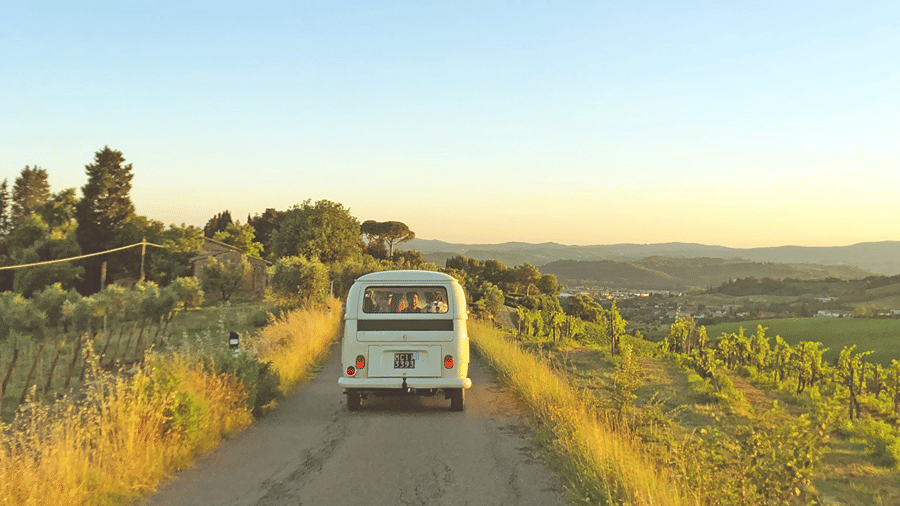 A van driving on a single lane road near a scenic landscape of green grass surrounded by trees, symbolising road trips from Delhi.