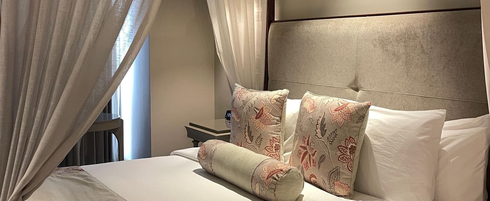 A side view of the four-poster bed in a Premium King Room at The Evren, Vagator. The bed is dressed with a bed runner and decorative pillows, and the white curtains are tied back, letting in natural light.