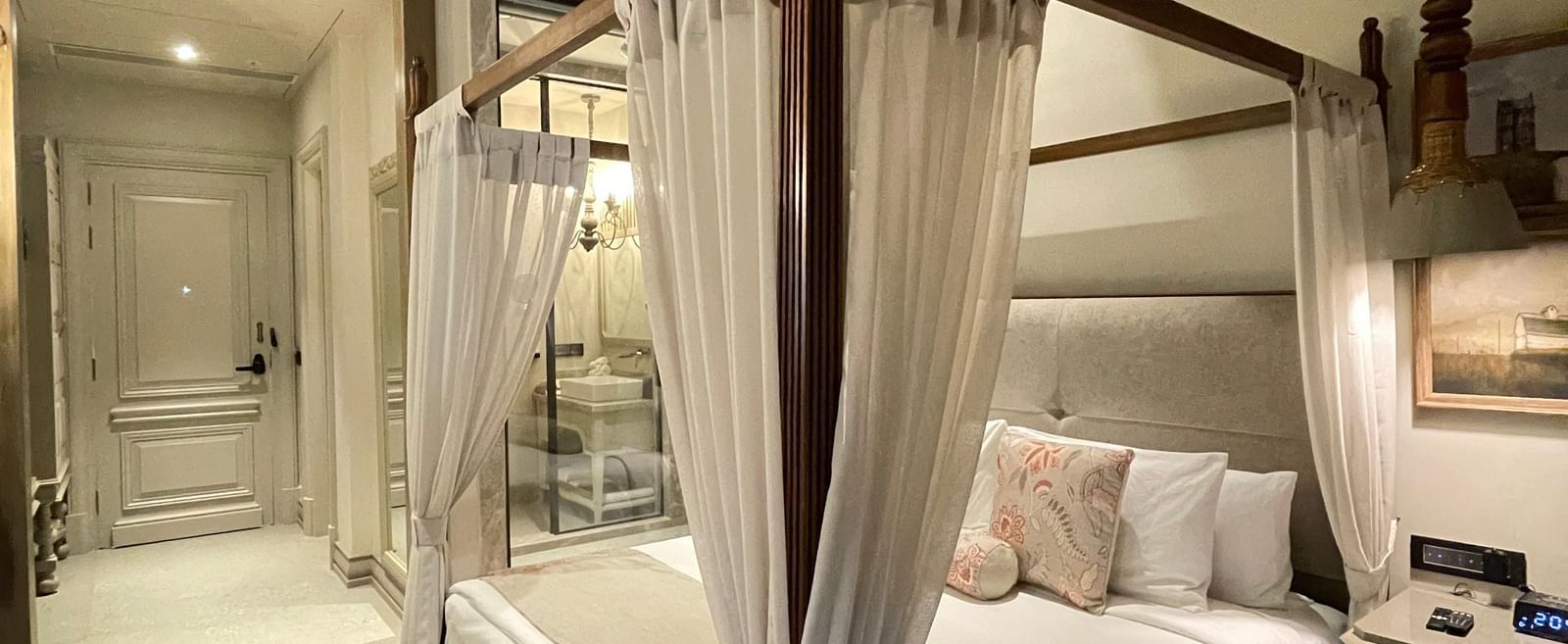 An elegant Premium King Room at The Evren, Vagator, featuring a four-poster bed with white curtains, a sloped wooden ceiling with a fan, and a view into the entryway. A patterned rug adds a touch of warmth to the space.