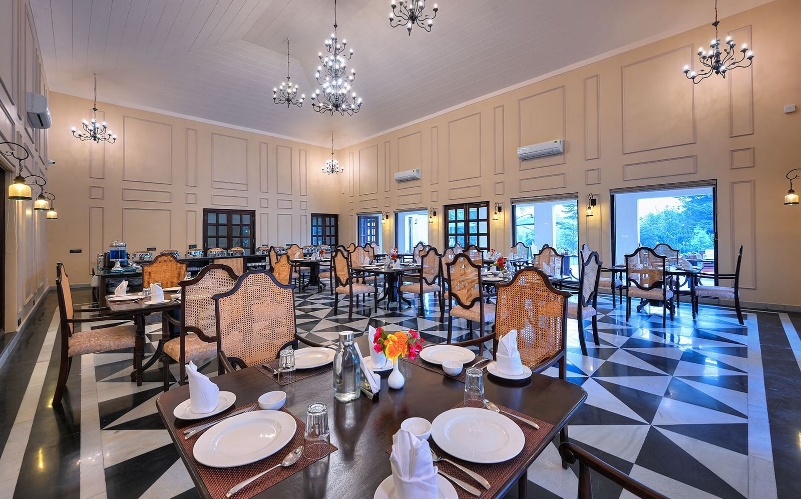 Spacious dining area featuring high ceilings, chandeliers, and classic wooden furniture. The black-and-white patterned flooring contrasts with the warm beige walls, complemented by large windows and elegant wall sconces - Singhvilas, Ranthambore