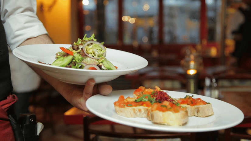 Waiter carrying two white plates, one with a fresh garden salad and the other with tomato bruschetta, in a cosy restaurant setting.