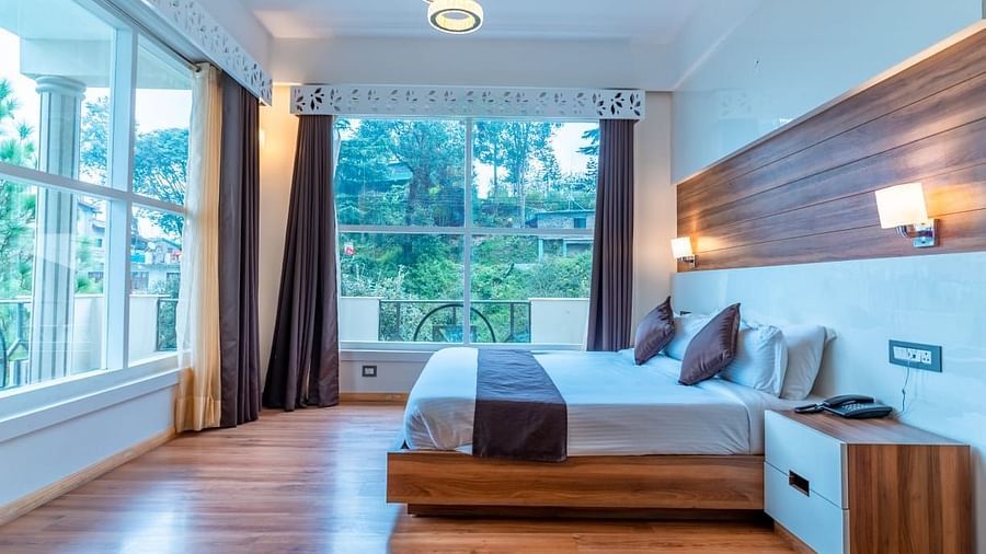 A bedroom with a white bed, a rug, and large windows, interior setting - Clarks Exotica Resort, Near Mukhteshwar.